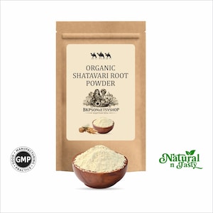 Puede incluir: Una bolsa de papel marr&oacute;n con la etiqueta "Organic Shatavari Root Powder". Un cuenco de madera lleno de polvo de color claro, con un cuenco m&aacute;s peque&ntilde;o y ra&iacute;ces tambi&eacute;n visibles. Las palabras "Natural n Tasty" est&aacute;n en verde.