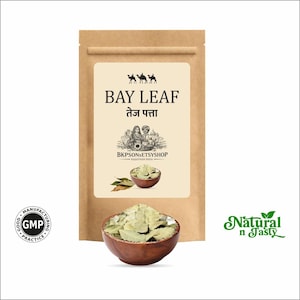 May include: A brown paper bag with a beige label featuring the text "BAY LEAF" and "तेज पत्ता". The label also includes an illustration of camels and people. A wooden bowl filled with bay leaves is in front of the bag.