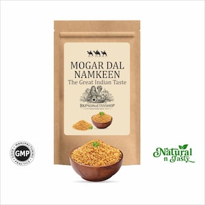 May include: A brown paper bag with a label that reads "MOGAR DAL NAMKEEN The Great Indian Taste." The label features illustrations of camels, people, and a bowl of food. A wooden bowl filled with the product is in the foreground.