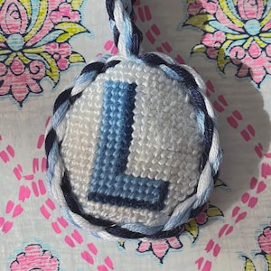 May include: A round, embroidered keychain featuring the letter "L" in shades of blue on a white background. The keychain is trimmed with a braided cord in blue and white. The background is a floral pattern.