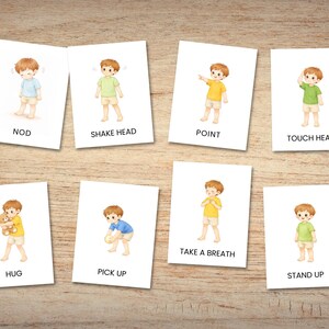May include: Eight flashcards with illustrations of a child demonstrating various actions. The cards include actions like "NOD," "SHAKE HEAD," "POINT," and "TOUCH HEAD." The child is wearing different colored shirts. The cards are on a wooden surface.