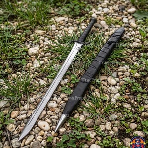 Puede incluir: Una espada de metal plateado con mango negro y guarda, acompañada de una vaina negra a juego. La espada presenta una hoja puntiaguda con una cresta central, mientras que la vaina tiene una punta de metal y un revestimiento de color negro.