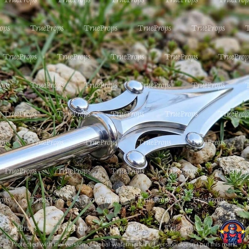 May include: A silver-colored metal spearhead featuring a pointed blade with a central ridge and decorative curved side prongs. The metal surface is polished and reflective, showcasing a smooth finish with rounded rivets connecting the components.