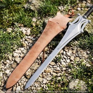 Puede incluir: Una espada de metal plateado con hoja puntiaguda y empu&ntilde;adura negra acanalada, que presenta una guarda decorativa con cuatro detalles circulares. La espada se acompa&ntilde;a de una funda de cuero color canela con correa y hebilla.