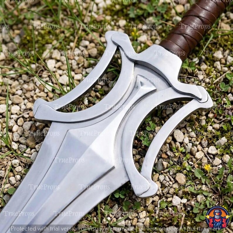 Puede incluir: Una espada de metal plateado con una guarda compleja y calada con brazos curvos y sim&eacute;tricos. La hoja tiene una cresta central y el mango est&aacute; envuelto en cuero marr&oacute;n. La espada est&aacute; colocada en diagonal dentro del encuadre.