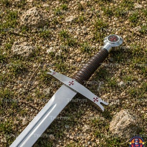 May include: A silver-toned metal sword featuring a dark brown ridged grip and a circular pommel. The crossguard and pommel are decorated with small red cross symbols. The blade is straight with a central fuller running down the length of the metal.