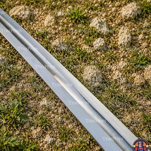 May include: A long, straight, double-edged metal sword blade with a central fuller running down the length of the steel. The blade features a polished, reflective surface that tapers toward a sharp point, showcasing a clean, metallic finish.