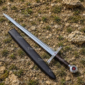 May include: A silver-toned metal sword with a double-edged blade, a brown leather-wrapped grip, and a circular pommel featuring a red cross emblem. The crossguard also displays small red cross details. A black scabbard lies beside the sword.