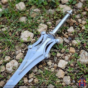 May include: A silver-colored metal dagger featuring a pointed blade and a decorative hilt with four circular rivets. The handle is cylindrical with a rounded pommel. The surface of the metal is reflective and smooth, showing a metallic finish.