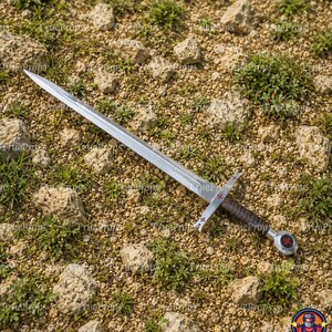 May include: A silver-toned metal sword featuring a pointed blade, a crossguard with small red cross emblems on each side, a dark brown wrapped handle, and a circular pommel with a red cross emblem centered on a black background.