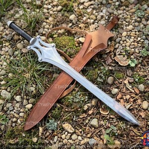 Puede incluir: Una espada de color plateado con mango negro y una guarda de metal compleja y sim&eacute;trica, acompa&ntilde;ada de una vaina de cuero color canela con punta afilada y una correa, colocada en diagonal dentro del encuadre.