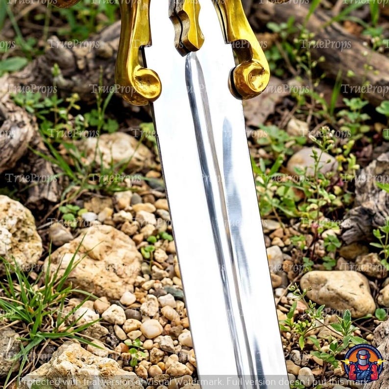 Puede incluir: Una hoja de espada de metal plateado con un surco central y un acabado pulido. La empu&ntilde;adura est&aacute; construida con guardas de metal dorado, ornamentadas y curvas, que se extienden hacia afuera desde la base de la hoja.