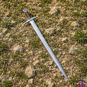 May include: A silver-toned metal sword featuring a long, double-edged blade with a central fuller. The crossguard and pommel are adorned with small red cross emblems. The handle is wrapped in dark brown cord, providing a textured grip.