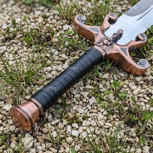 Puede incluir: Una espada con una guarda de metal color cobre que presenta un emblema de calavera central y gavilanes curvos. El mango est&aacute; envuelto en material negro y termina en un pomo a juego color cobre. La hoja es plateada con un patr&oacute;n ondulado visible.
