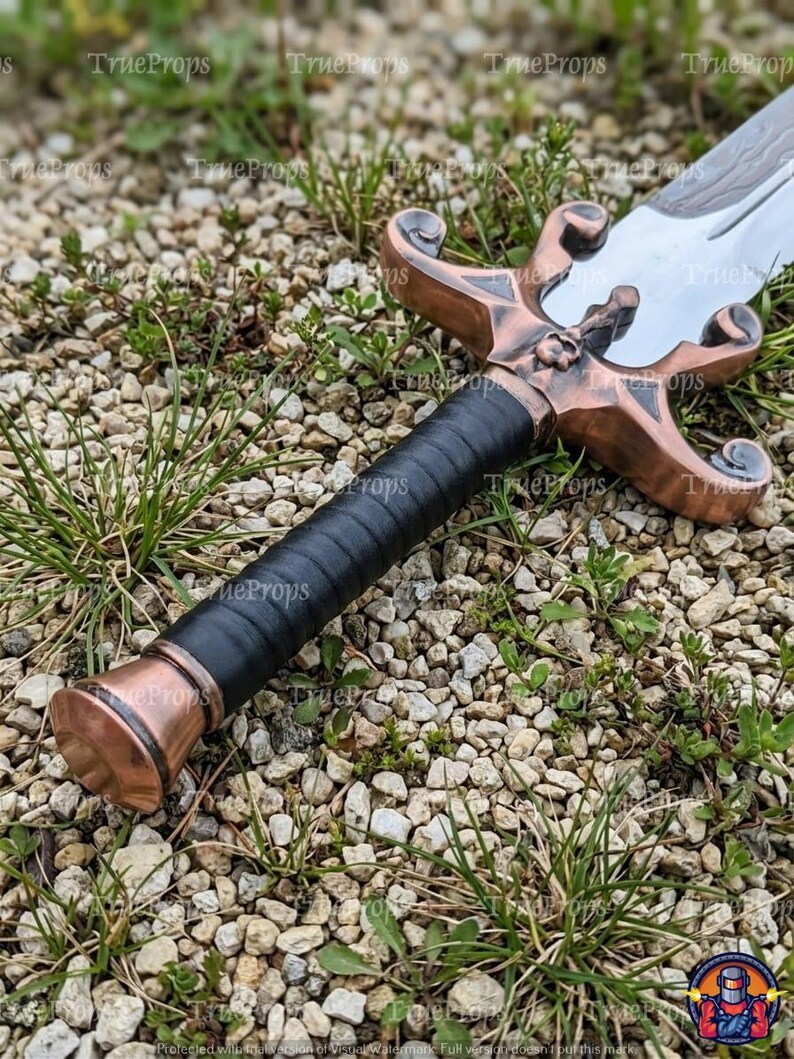 Puede incluir: Una espada con una guarda de metal color cobre que presenta un emblema de calavera central y gavilanes curvos. El mango est&aacute; envuelto en material negro y termina en un pomo a juego color cobre. La hoja es plateada con un patr&oacute;n ondulado visible.