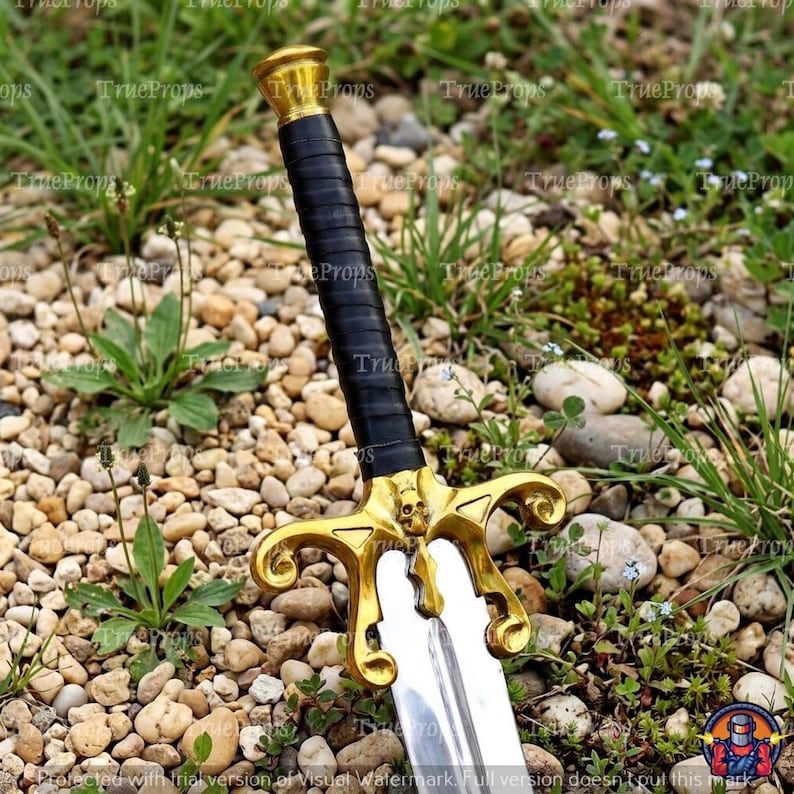 Puede incluir: Una espada con hoja de plata pulida, empu&ntilde;adura envuelta en negro y una guarda de color dorado con un peque&ntilde;o detalle de calavera en el centro. El pomo es una tapa sencilla de color dorado y la guarda tiene extremos curvos y ornamentados.