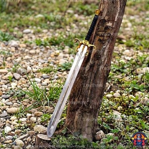 Puede incluir: Una espada larga con hoja de metal plateado, una guarda dorada con detalles ornamentales y una empu&ntilde;adura negra texturizada. La espada est&aacute; colocada verticalmente contra un poste de madera rugoso y desgastado al aire libre.
