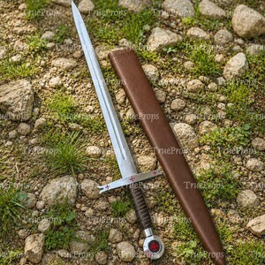 May include: A silver-toned metal sword with a double-edged blade, a crossguard featuring red cross emblems, and a dark brown leather-wrapped grip. The sword includes a matching brown leather scabbard with a rounded pommel displaying a red cross.