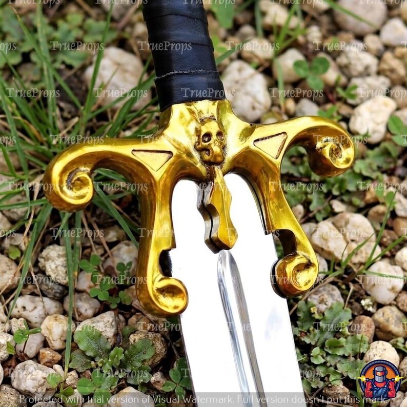 Puede incluir: Una espada con hoja de plata met&aacute;lica y una guarda de color dorado que presenta un emblema de calavera central. El mango est&aacute; envuelto en material negro. La guarda tiene extremos curvos y ornamentados que se extienden hacia afuera.