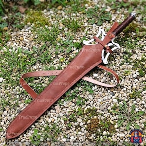 Puede incluir: Una espada con empu&ntilde;adura envuelta en cuero marr&oacute;n oscuro y una guarda de metal plateado con un dise&ntilde;o calado intrincado. La hoja est&aacute; enfundada en una vaina de cuero marr&oacute;n a juego con una correa de cuero larga y ajustable.