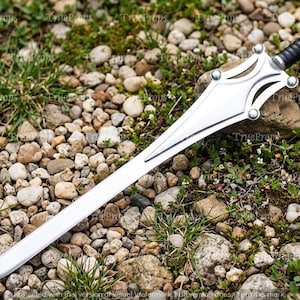 Puede incluir: Una espada de color plateado con una hoja puntiaguda y una guarda de metal estilizada con cuatro acentos circulares. El mango es negro con una textura acanalada y un pomo met&aacute;lico en el extremo. La hoja es lisa y termina en punta.