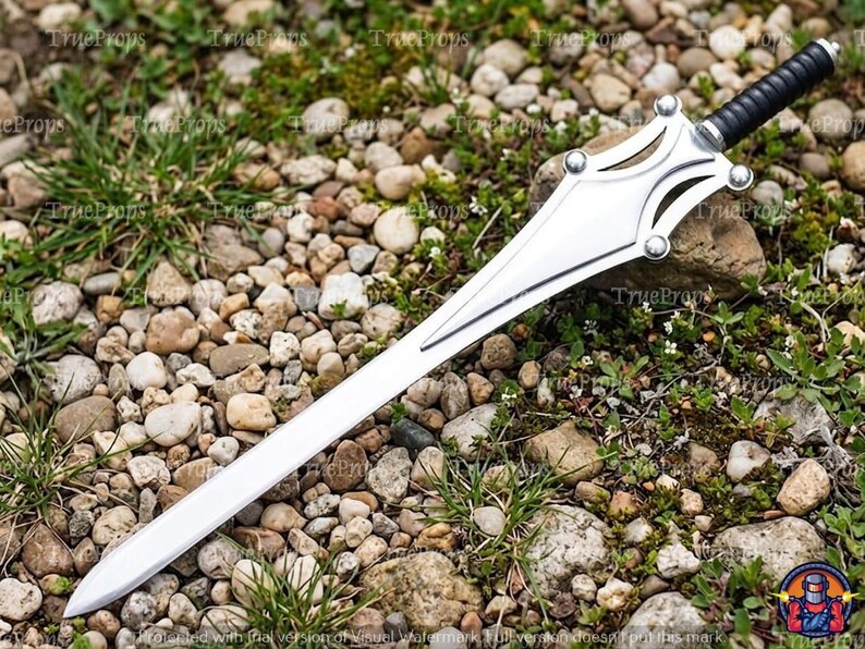 Puede incluir: Una espada de color plateado con una hoja puntiaguda y una guarda de metal estilizada con cuatro acentos circulares. El mango es negro con una textura acanalada y un pomo met&aacute;lico en el extremo. La hoja es lisa y termina en punta.