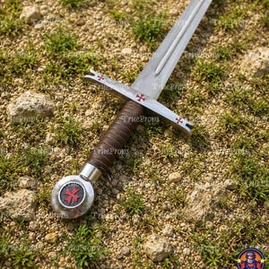 May include: A metal sword featuring a silver blade, a brown leather-wrapped grip, and a circular pommel with a red cross emblem. The crossguard is decorated with three small red crosses, and the pommel displays a prominent red Maltese cross.