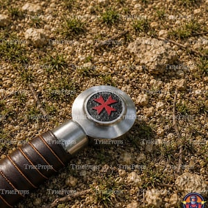 May include: A sword pommel featuring a circular silver metal design with a textured black center and a red Maltese cross emblem. The handle is wrapped in dark brown leather with visible orange stitching, showing a metallic finish.