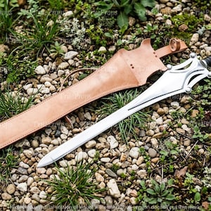Puede incluir: Una espada de metal plateado con hoja puntiaguda, guarda decorativa y mango envuelto en negro, acompa&ntilde;ada de una vaina de cuero color canela con bordes cosidos y una correa de sujeci&oacute;n, sobre un lecho de peque&ntilde;as piedras y hierba.