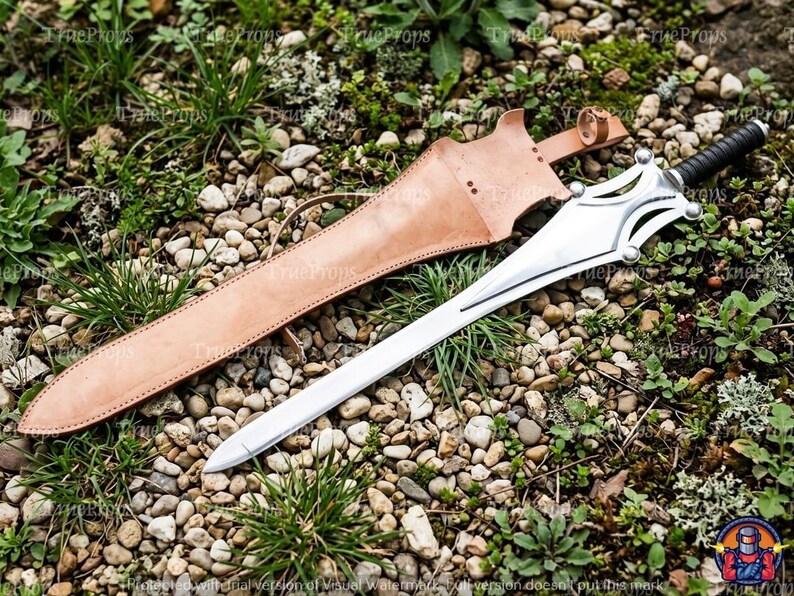 Puede incluir: Una espada de metal plateado con hoja puntiaguda, guarda decorativa y mango envuelto en negro, acompa&ntilde;ada de una vaina de cuero color canela con bordes cosidos y una correa de sujeci&oacute;n, sobre un lecho de peque&ntilde;as piedras y hierba.