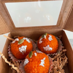 Puede incluir: Tres dulces redondos de color naranja, con acentos de hojas de plata y una semilla verde en la parte superior, presentados en una caja marrón con una ventana transparente. Los dulces están en copas de papel decorativas.