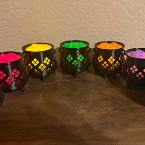May include: A collection of seven black plastic cauldrons arranged in a semi-circle. Each cauldron features a diamond-shaped lattice cutout pattern and contains a brightly colored insert in red, pink, yellow, green, orange, purple, and blue.