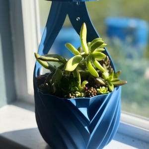 Op de afbeelding: Een blauwe hangende plantenbak met een geometrisch ontwerp, gevuld met groene vetplanten. De plantenbak is opgehangen aan een gebogen bovenkant, wat een moderne, decoratieve uitstraling creëert. De planten bloeien en voegen een vleugje natuur toe.