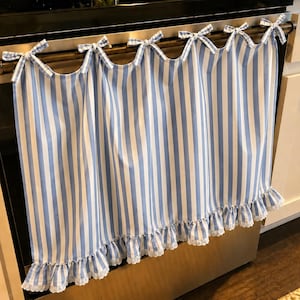 Puede incluir: Una cortina de rayas verticales azul claro y blancas colgada del tirador de una estufa. La cortina presenta cinco lazos de tela en el borde superior y un dobladillo con volantes adornado con encaje blanco. La tela tiene un patrón uniforme.