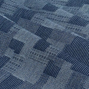 May include: Close-up of a textured fabric with a patchwork design. The fabric features a mix of geometric patterns in shades of blue, including stripes, dots, and squares. The fabric has a slightly wrinkled appearance, suggesting it is soft and pliable.