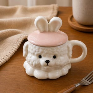 Könnte beinhalten: Eine weiße Keramiktasse in Hundeform mit rosa Deckel und Hasenohrengriff. Die Tasse hat einen runden Griff und schwarze Augen. Die Tasse steht auf einer Holzoberfläche.