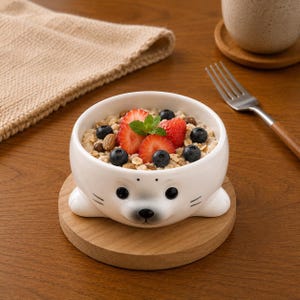 Può includere: Una ciotola in ceramica bianca a forma di foca, riempita con farina d'avena, fragole, mirtilli e noci. La ciotola è appoggiata su un sottobicchiere di legno. Una forchetta e una tazza sono sullo sfondo.