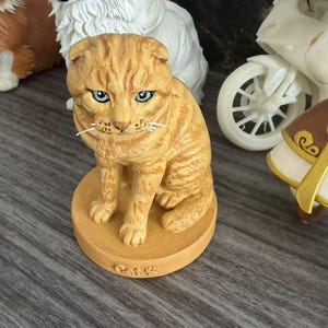 May include: A small orange tabby cat figurine with folded ears and blue eyes, sitting on a round base with the word Cat embossed on the front. The cat has detailed fur texture and white whiskers, positioned in front of a white dog figurine.