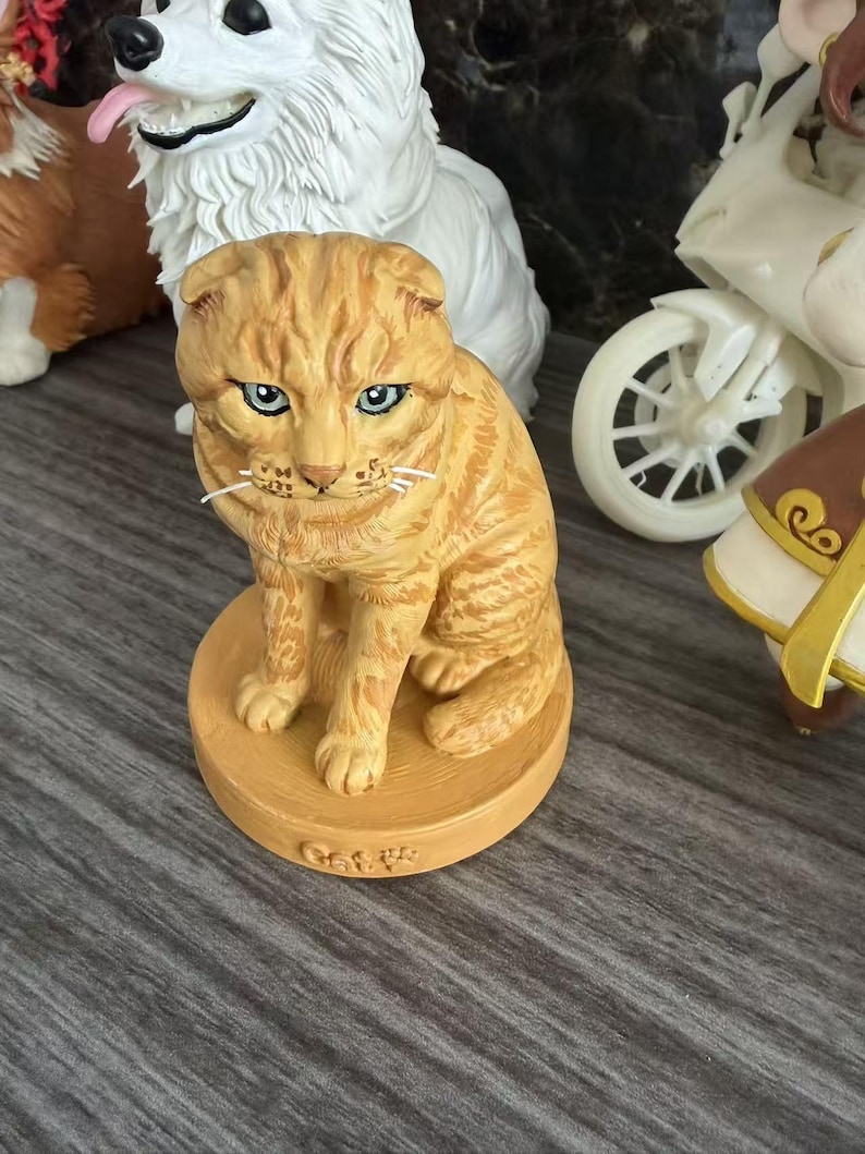 May include: A small orange tabby cat figurine with folded ears and blue eyes, sitting on a round base with the word Cat embossed on the front. The cat has detailed fur texture and white whiskers, positioned in front of a white dog figurine.