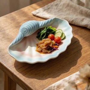 Könnte beinhalten: Eine Keramik-Servierschale in Form einer großen Muschel mit einem hellblauen und weißen Farbverlauf. Die Schale enthält eine kleine Portion gekochtes Blattgemüse, Gurkenscheiben, Kirschtomaten und eine herzhafte Pfannengemüsemischung.