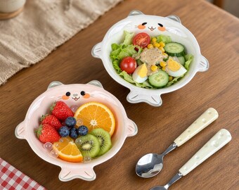 May include: Two ceramic bowls shaped like cats, one white and one light pink, each featuring a cartoon face. The white bowl contains a salad with lettuce, tomatoes, cucumbers, corn, and egg. The pink bowl holds strawberries, orange slices, blueberries, and kiwi.