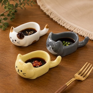 May include: Three ceramic cat-shaped sauce dishes in white, gray, and yellow, each filled with a dark sauce and garnishes. The dishes have a tail handle and cat face details. A gold fork with a brown handle is in the foreground.