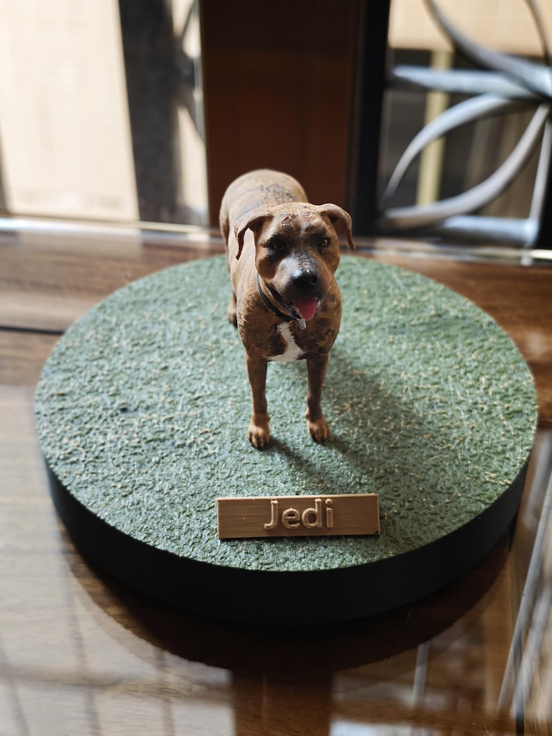 May include: A brown and white brindle dog figurine standing on a circular green textured base. The dog has a black collar and a pink tongue visible. A rectangular plaque attached to the front of the base features the name Jedi in raised letters.