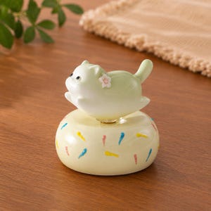 Pode incluir: Uma figura de cerâmica de um gato verde claro com uma flor rosa na lateral, pousada sobre uma base amarela em forma de donut. A base é decorada com traços de tinta coloridos e abstratos. A figura do gato tem olhos pretos e cauda levantada.