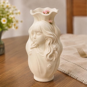 Könnte beinhalten: Eine mattweiße Keramikvase mit einem skulpturalen Relief einer Person mit langem, welligem Haar und Pony. Die Vase hat einen gewellten, geschwungenen Rand und ist mit kleinen geprägten Blumenmotiven verziert, mit winzigen rosa Blüten im Inneren.
