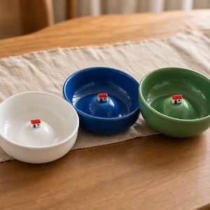 May include: Three ceramic bowls in white, blue, and green, each featuring a small, raised mound in the centre topped with a miniature white house figurine that has a red roof and a blue door, arranged in a row on a textured surface.