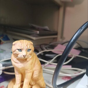May include: A small, orange tabby cat figurine with folded ears and blue eyes, sitting on a round, tan base. The base features the word "Cat" embossed on the front. The figurine is rendered in a realistic style with detailed fur texture.