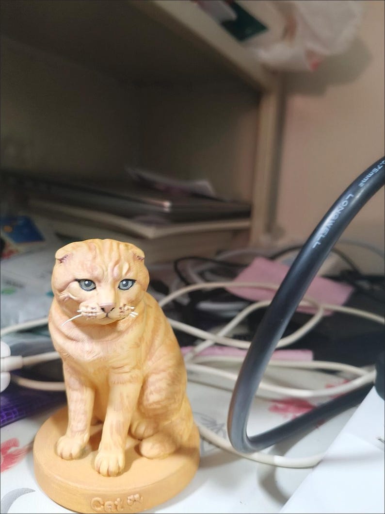 May include: A small, orange tabby cat figurine with folded ears and blue eyes, sitting on a round, tan base. The base features the word "Cat" embossed on the front. The figurine is rendered in a realistic style with detailed fur texture.
