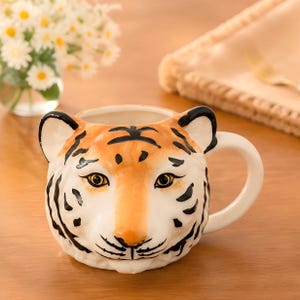 Könnte beinhalten: Ein Keramikbecher in Form eines Tigerkopfes mit orange-weißem Fellmuster, schwarzen Streifen, detaillierten Augen und einem geschwungenen Henkel auf der rechten Seite. Der Becher hat eine weite Öffnung und ein geformtes Gesicht.