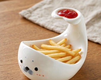 May include: A white ceramic bowl shaped like a seal, filled with golden-brown french fries. The bowl features a small, elevated side compartment containing red ketchup, and a painted face with black eyes, a gray nose, and pink cheeks.